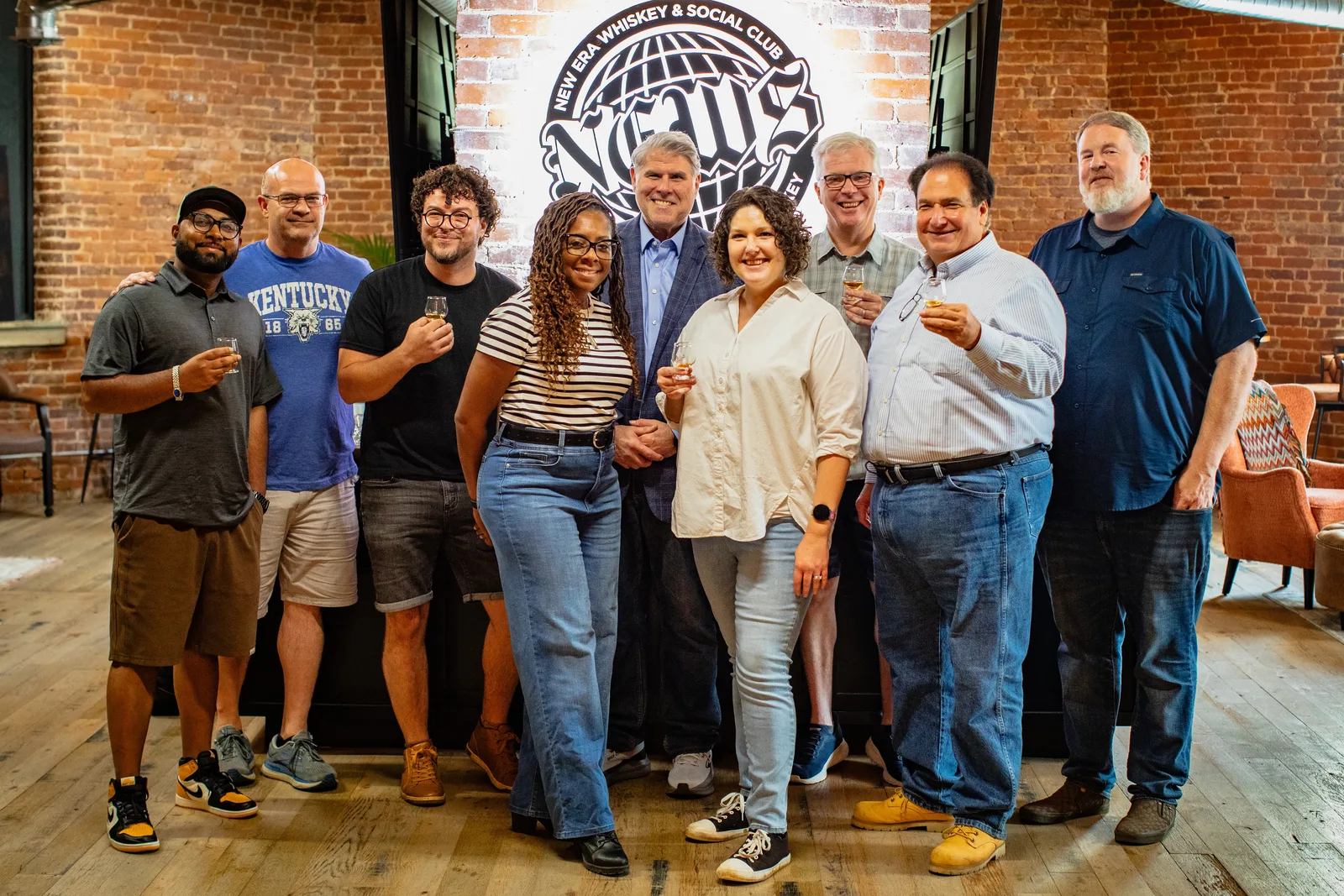 The nine Batch #1 tastemakers standing together in front of the New Era Whiskey & Social Club sign, most raising a tasting glass